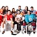 A group of eight young adults, each dressed in different sports uniforms, are posed together smiling. They hold various sports equipment including a soccer ball, volleyball, basketball, football, tennis racket, and table tennis paddle. The clothing includes jerseys and gear for soccer, hockey, football, and other sports. The background is plain white, emphasizing the colorful attire of the group.