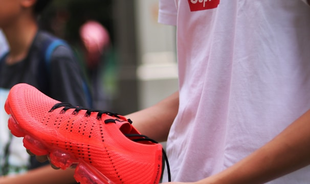 A person holding a vibrant orange sports shoe with translucent soles, standing next to someone else. The person is wearing a white t-shirt with a distinct logo in red and black. The background is muted, suggesting an urban environment.