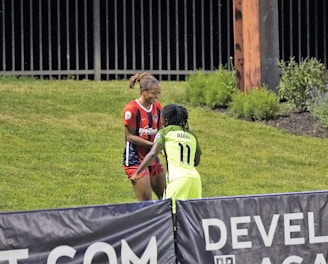 Two individuals are interacting on a grassy field. One is wearing a red and blue sports outfit with 'ProChain' visible, and the other is in a bright yellow uniform with the name 'Addo' and the number 11. They appear to be playing a sport, possibly soccer, as they are near a marked boundary. A fence and some vegetation are visible in the background.