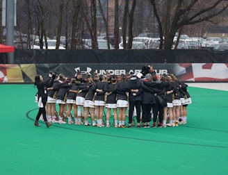 A group of athletes is gathered in a huddle on a sports field, wearing matching uniforms and gear. The ground is green, and there is signage with a brand logo in the background. Trees and parked cars can be seen behind a fence surrounding the field. A person on the left side is capturing the moment with a camera.