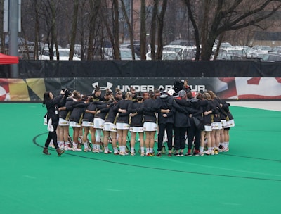 A group of athletes is gathered in a huddle on a sports field, wearing matching uniforms and gear. The ground is green, and there is signage with a brand logo in the background. Trees and parked cars can be seen behind a fence surrounding the field. A person on the left side is capturing the moment with a camera.