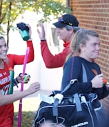 A group of athletes in sports attire appears to be in a joyful mood. One person is holding a field hockey stick and wearing a headband, while another carries a sports bag. They are gathered near a brick wall with trees in the background, sharing a light moment, likely after a sports event.