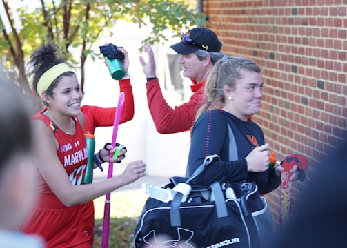 A group of athletes in sports attire appears to be in a joyful mood. One person is holding a field hockey stick and wearing a headband, while another carries a sports bag. They are gathered near a brick wall with trees in the background, sharing a light moment, likely after a sports event.