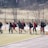 A group of individuals dressed in athletic clothing walk across a sports field. Many of them are carrying sports bags and some hold lacrosse sticks. They appear to be part of a team, wearing matching jackets and sneakers. The field is bordered by a fence and some trees in the background.