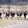 A group of individuals dressed in athletic clothing walk across a sports field. Many of them are carrying sports bags and some hold lacrosse sticks. They appear to be part of a team, wearing matching jackets and sneakers. The field is bordered by a fence and some trees in the background.