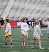 A group of young women wearing athletic gear are on a sports field. They are dressed in white jerseys with yellow shorts, featuring a distinctive design at the hem. The field has a green turf surface and the background shows empty bleachers. Some additional figures are visible in the distance, possibly referees or other players.