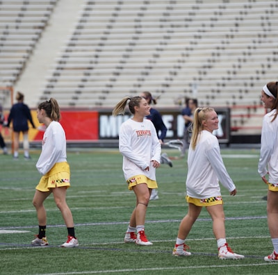 A group of young women wearing athletic gear are on a sports field. They are dressed in white jerseys with yellow shorts, featuring a distinctive design at the hem. The field has a green turf surface and the background shows empty bleachers. Some additional figures are visible in the distance, possibly referees or other players.