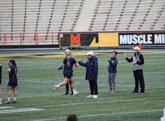 Several people are on a sports field with an empty bleacher in the background. One person is high-fiving a coach, while others stand nearby. They are wearing athletic clothing, hinting at a team sport environment. A sign with the text 'Muscle Milk' is visible in the background.