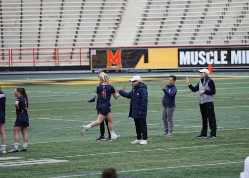 Several people are on a sports field with an empty bleacher in the background. One person is high-fiving a coach, while others stand nearby. They are wearing athletic clothing, hinting at a team sport environment. A sign with the text 'Muscle Milk' is visible in the background.