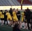 A group of athletes wearing yellow sports uniforms are standing on a sports field. Some are holding lacrosse sticks, and they appear to be interacting casually. Several people are dressed in black coats. In the background, there is a bright sign with text and parts of a stadium are visible.