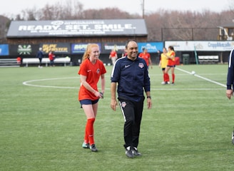 A group of people are on a soccer field, with some wearing sports attire. In the foreground, a person in red sports clothing is walking alongside someone in a dark tracksuit. Other people are visible in the background, some wearing yellow jerseys. A building with advertising banners, including a prominent sign reading 'Ready, Set, Smile,' is in the background.