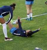 A group of athletes in sportswear are on a green soccer field. Two individuals are interacting closely; one helps the other who is lying on the grass. Nearby, other athletes are stretching.