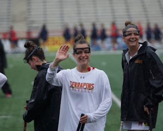 Several athletes in sports attire are on an outdoor sports field, interacting and smiling. One individual in the foreground waves with a friendly expression, while another stands beside her, also smiling. They wear protective goggles and gear associated with lacrosse. The background features indistinct figures in dark clothing against a backdrop of stadium seating.