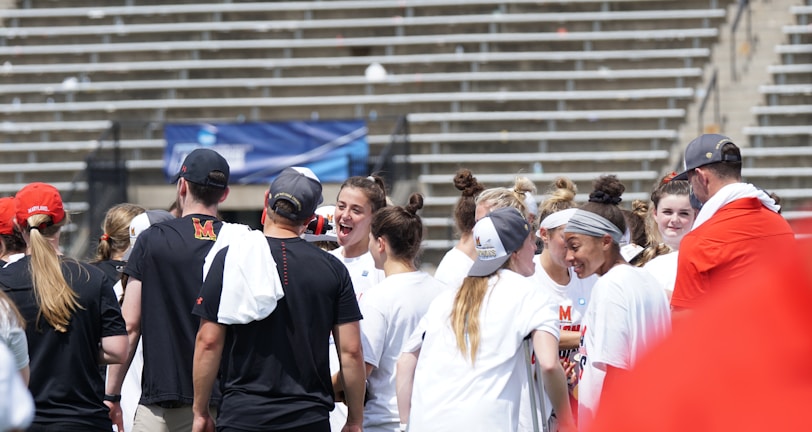 A group of athletes in white shirts with black and red accents are gathered together, engaged in conversation and smiling. They appear to be on a sports field with empty bleachers in the background. The setting is outdoors, and it seems sunny. Some individuals are wearing caps or headbands.