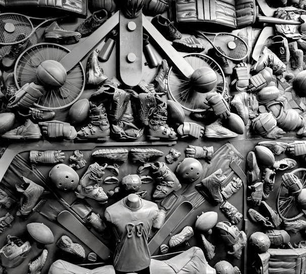 A complex arrangement of various sports equipment and attire, including tennis rackets, soccer balls, basketballs, helmets, shoes, gloves, and a mannequin torso wearing a T-shirt. The items are layered and artistically displayed in a monochrome color scheme, creating an intricate texture and visual pattern.
