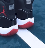 A pair of black and white sneakers with red soles placed on a blue outdoor sports court. The shoes feature a distinct red silhouette logo on the back. The court shows a white line separating two sections.