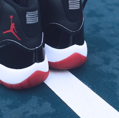 A pair of black and white sneakers with red soles placed on a blue outdoor sports court. The shoes feature a distinct red silhouette logo on the back. The court shows a white line separating two sections.