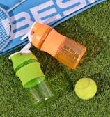 Two colorful plastic bottles, one orange and one green, rest on artificial grass next to a yellow tennis ball and a badminton racket with a blue sports bag partially visible.