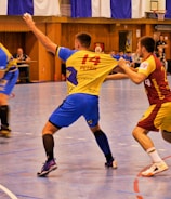 A dynamic indoor sports scene with players in motion on a court. Two athletes in blue and yellow uniforms are engaged in an intense handball game, with one player gripping the other's jersey. The background shows other players, several seated people, and advertisements on the walls.