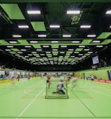 A sports hall with bright green flooring and ceiling panels. There is a game in progress with players moving quickly, suggesting a fast-paced sport like floorball or indoor hockey. Advertising banners line the walls, and a scoreboard is visible in the background.