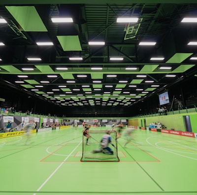 A sports hall with bright green flooring and ceiling panels. There is a game in progress with players moving quickly, suggesting a fast-paced sport like floorball or indoor hockey. Advertising banners line the walls, and a scoreboard is visible in the background.