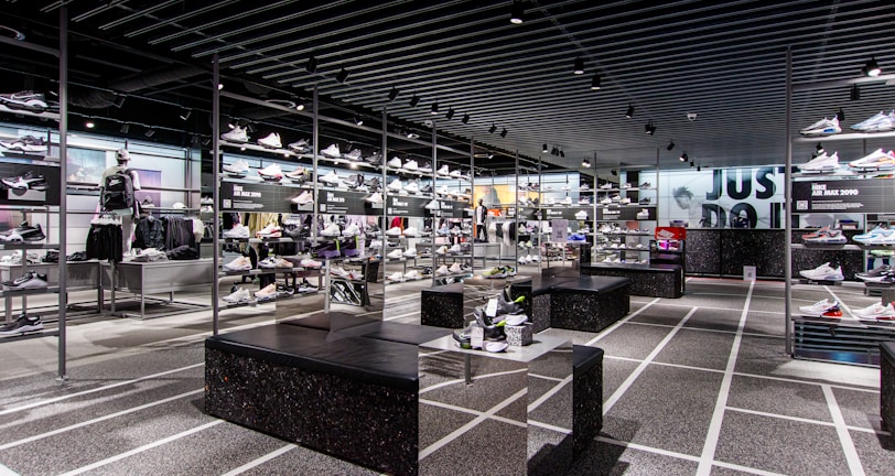 A modern sneaker store with sleek black shelving displaying various athletic shoes and sports apparel. Central seating areas and spotlights provide a contemporary retail ambiance. The signage prominently features a famous sportswear slogan in the background.