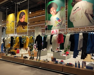 A retail store showcases clothing and sportswear on mannequins and hangers, with large vibrant posters featuring children in sports attire above. The interior displays bright garments and accessories on shelves and racks.