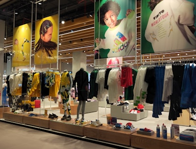 A retail store showcases clothing and sportswear on mannequins and hangers, with large vibrant posters featuring children in sports attire above. The interior displays bright garments and accessories on shelves and racks.