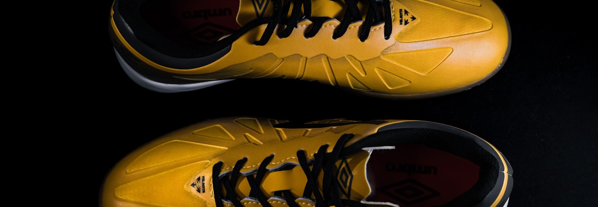 A pair of yellow sports shoes with black laces is placed against a dark background. The design is sleek, with angular patterns on the surface. The inside of the shoes appears to have a red lining with a brand logo visible.