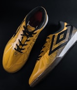 A pair of yellow and black sports shoes displayed against a dark background. The shoes feature a bold geometric pattern and are designed for athletic performance.