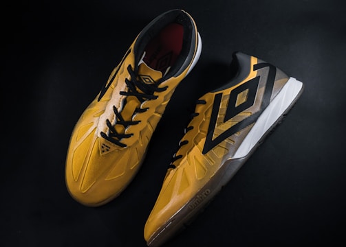 A pair of yellow and black sports shoes displayed against a dark background. The shoes feature a bold geometric pattern and are designed for athletic performance.