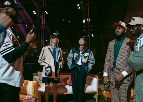 A group of people are gathered indoors, wearing stylish casual and sports-themed outfits, including jackets and caps. Some are standing while one appears to be taking a photo. The environment has a warm, wooden interior with visible beams and spotlights, and a cozy arrangement of furniture including chairs and a sofa.